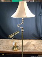 Full view of tall brass floor lamp with beige fabric bell-shaped shade set against a dark background on a wooden surface.
