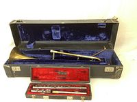 Full view of the open black case with a brass slide trombone inside, lined with blue velvet, and a second red-lined case containing parts of a silver-colored flute.