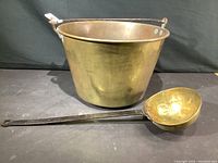 Full view of antique copper pail with iron handle alongside hammered copper ladle on a flat surface