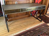 Front view of desk showing greyed oak top, three brass-clad drawer fronts, black metal X-shaped legs