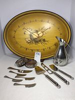Overall view of the lot showing the serving tray with musical instruments design, pewter pitcher, wine coasters, and cheese cutlery laid out.