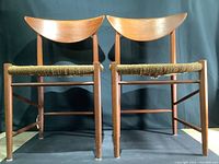 Front view of two Danish modern teak dining chairs with curved bent backrests and woven rush seats, one seat visibly worn.