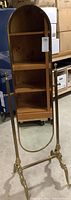 Full view of the standing mirror showing its height, brass frame, and wooden shelving in the background.