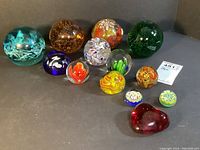 Group of 13 assorted glass paperweights displayed on gray surface in two rows