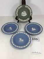 Photo of entire lot showing one sage green ash receiver plate on stand and three blue coaster dishes in front, clearly showing white relief designs of mythological figures and floral garlands.