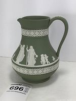 Front view of sage green Wedgwood Jasperware pitcher showing white classical reliefs of figures and cherubs and floral garland band.