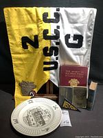 Overview photo showing yellow and white USCC Unit 2G garrison flag, challenge coin, cadet device, four military-related books, display plate with military insignia and Citadel illustration, and West Point pennant.