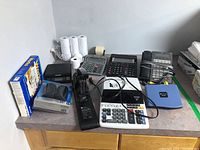 Full view of the lot items on a table including two Casio adding machines, one Sharp adding machine, NEC phone, Dynex cable wrap, stapler, Microsoft Works software, Dymo scale, and multiple paper rolls.