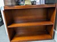 Front view of wooden bookcase showing two shelves, medium wood tone, surface scratches and scuffs visible.
