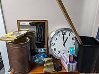 Photo showing round clock, framed mirror, metal trash can, black plastic wastebasket, wooden yardstick, box of bandaids and face masks, small blue glass bowl, metal bookend, pencil basket, and set of crossword puzzle books.