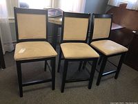 Three counter height chairs with beige upholstered seats and backs, dark wooden frames, positioned in a row indoors