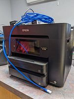 Front and side view of the black Epson Workforce Pro WF-4740 multifunction printer with blue Ethernet cable draped over it.