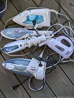 Photo showing two clothing irons (Black & Decker First Impressions and T-FAL Ultra Glide) and three handheld steamers (Conair, Steam Buddy, Epica) with cords tangled, items placed on a wooden surface.