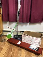 Photo showing Shark vacuum cleaner, green broom, white dustpan, cardboard box, red wooden wall shelf laid on floor, two yard sticks leaning on box, and packaged outlet covers lying on shelf.