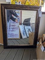 Full view of a large rectangular wall mirror with a wide dark brown textured frame. Mirror surface visibly scratched and reflects other framed art pieces lying nearby.