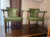 Pair of green vinyl upholstered wooden arm office chairs with nail head trim shown side by side on a wooden floor near a window and curtain.