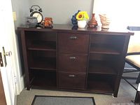 Front view of dark wood office cabinet showing three open shelves on each side and three file drawers in the middle, including decorative items on top and visible surface wear.
