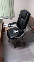 Black padded vinyl office chair with armrests, placed on a protective floor mat near a desk, showing wear on arms.
