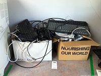 Photo showing two boxes filled with various black keyboards and cables; cords visible hanging out of boxes