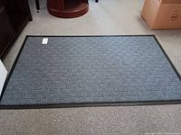 Rug mat lying flat on carpeted floor, showing overall rectangular shape and black rubber border around the edges.