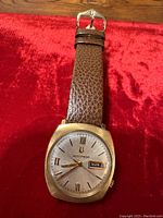 Front view of watch showing cushion-shaped gold case, silver dial with Roman numerals and day-date window