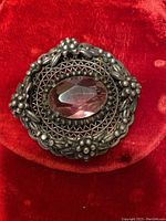 Front view of antique silver brooch with large oval faceted purple amethyst stone, set in detailed floral and lattice pattern.