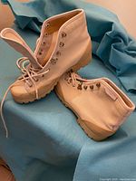 Pair of white canvas hi-top shoes with beige rubber sole and metal lace eyelets on a blue fabric background.