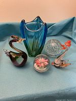 Photo of a medium blue-green glass vase with flared open top, two glass bird figurines with orange beaks, and three round glass paperweights on a light blue fabric background.