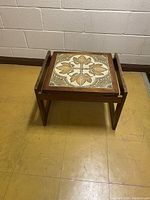 Full view of wooden side table with tile inlay top against white wall and yellow floor.