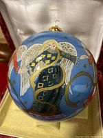 Front view of blue round ornament showing angel with yellow hair, white wings, blue and gold robes, holding a star and crescent shapes.