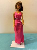 Full length image of Barbie doll standing on white stand with pink long dress and one visible high heel shoe.