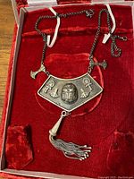 Front view displaying the necklace panel with raised Egyptian symbols: scarab, ankhs, lotus flowers, and hanging tassel detail on red velvet backing.