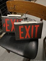 Two vintage electric exit signs on a chair, showing front and side views, with red EXIT letters on black frames and attached electrical wiring.