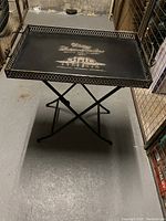 Black metal folding serving table shown upright with tray top and decorative pierced edge.