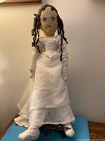 Full body of vintage handmade cloth doll standing against a wall, dressed in a long white dress with veil and silk shoes