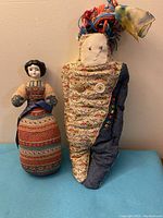 Two dolls shown standing: one tall cloth doll with hand embroidery and buttons, one smaller ceramic-headed doll in patterned dress.