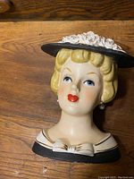 Photo of vintage ceramic head vase showing woman's face with pearl earrings, blonde hair, and black hat with white flower details.