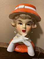 Front angled view of the head vase bust showing the woman's face, orange hat with white band and buckle, pearl earrings, pearl necklace, and white gloves.