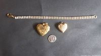 Photo showing the diamond-effect bracelet with gold backing laid horizontally above two gold-colored heart lockets (one hand engraved, the other smooth), with a quarter for size comparison.