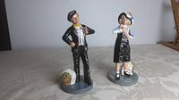 Front view of both Royal Doulton Pearly Girl and Pearly Boy figurines standing on grey circular bases with added accessories like basket