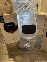 Photo showing the clear stemmed wine glass with a rectangular black chalkboard label on the front, next to its box with 'Pinot noir' depiction.