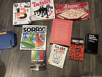 Top view of multiple board games arranged on floor including Scrabble, Twister, Sorry!, Balderdash, Cards Against Humanity, Mad Libs, You Are a Liar, Fake News, and Battleship.
