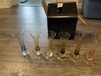 Six Bombay stemmed shot glasses with colored stems arranged in a row in front of the box on a wood floor.