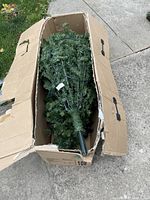 Artificial Christmas tree inside a worn cardboard box, branches tightly packed with visible tags and wiring.