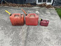 Wide angle photo showing all three red gasoline containers placed on concrete surface outdoors, showcasing size variation and condition.