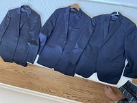 Three men's suit jackets in dark colors laid out on white surface showing front view.