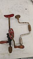 Two vintage hand drills laid flat showing red eggbeater style crank drill and smaller crank drill with round metal guard.