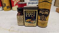 Photo showing the small amber glass bottle of La Perle Clock Hammel Riglander oil, Green's Gravy Salt tin, and Pure Brand oil can together.