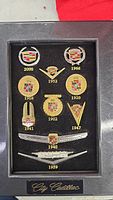 Display of a black velvet case containing 12 Cadillac emblem pins from various years including 1902 to 2000, arranged in neat rows with City Cadillac inscription.