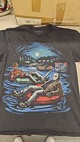 Front of black T-shirt showing graphic of Freddy Krueger and Jason Voorhees on inflatable tubes on water near Camp Crystal Lake sign at night with moon.